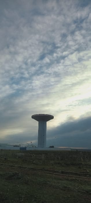 A vertical shot features a concrete tower against a backdrop of a cloudy, partly sunny sky. The tower has a distinct, flared top. Adjacent to the tower is a patch of green grass and a paved area. The sky is filled with wispy clouds, with the sun peeking through, creating a bright, diffused light. The overall image has a serene and somewhat industrial feel.