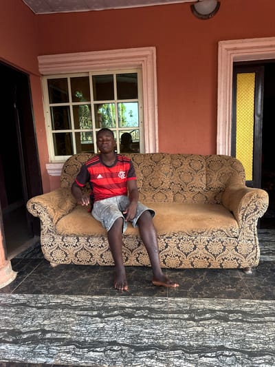 A man in a red and black soccer jersey and gray shorts sprawls on a gold patterned sofa. He faces the viewer with eyes closed. The sofa is placed on dark marble-look tile outside a salmon-colored building with white trim. A multi-pane window reflects trees. A small light fixture is affixed to the wall. Another window with a gold-toned panel is above.