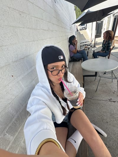 The image depicts an outdoor cafe patio located at 1417 C in Glendale, United States, under bright daylight. A young woman is seated in the foreground, positioned on a wooden bench and leaning against a white textured brick wall. She wears a black baseball cap featuring an MLB logo, tortoiseshell-rimmed glasses, a white hooded zip-up sweatshirt, a black tank top, denim shorts, white socks with a black Nike swoosh, and black footwear. Her dark hair is styled in two braids, and she wears a gold-colored bracelet on her left wrist and a necklace with two small pendants. She holds a clear plastic cup containing a dark beverage with a white sleeve and a pink straw to her mouth, adopting a pouting expression towards the camera.
In the midground, a second woman with dark skin and dark hair is seated further down the same wooden bench, wearing a purple top and blue jeans. Her left hand is raised towards her mouth. To her right, a third woman with curly reddish-brown hair is seated at a round, light-colored table with a dark metal chair, dressed in a plaid shirt and blue jeans, looking towards the second woman.
The patio area includes two round tables with dark metal bases and two black patio umbrellas. A white building with a glass storefront and the address "1417 C" is visible in the background. A concrete or asphalt ground extends into a street with a gentle incline, where a white vehicle is partially visible in the distance. Green foliage appears on a hillside in the far background.