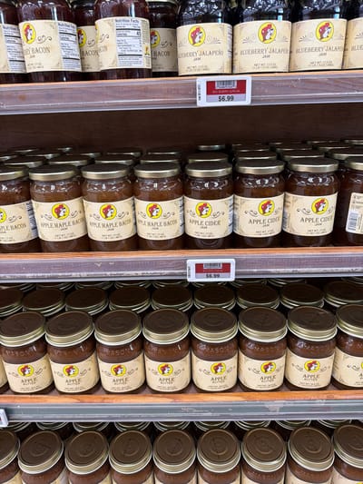 This media file is a close-up, eye-level shot of shelves filled with jars of preserves, likely in a grocery store in Katy, United States. The shelves are made of wood, and a clear plastic divider runs vertically down the center of the frame, separating two distinct rows of products.

On the left side of the divider, the jars contain jams and jellies. The labels prominently feature a cartoon beaver character and text indicating flavors such as "BLUEBERRY JALAPEÑO JAM" and "APPLE MAPLE BACON JAM." Some of these labels also display nutritional information and weight specifications in ounces and grams. A price tag for "$6.99" is visible on one of the "BLUEBERRY JALAPEÑO JAM" jars.

On the right side of the divider, the jars contain "APPLE CIDER JAM" and "APPLE BUTTER." These jars have similar labeling styles, including the beaver character, but with different flavor names. The lids of these jars are a metallic color, some with embossed lettering that appears to say "MASON."

The lighting is bright and even, suggesting an indoor setting with artificial lights. The overall impression is one of abundant product display, typical of a retail environment. The focus is entirely on the products, with no people or other extraneous elements visible.