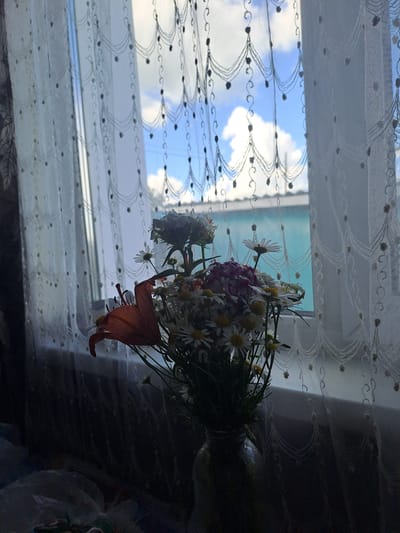 The image depicts an indoor scene with a bouquet of flowers in a vase placed in front of a window with white lace curtains. Outside the window, a blue sky with white clouds and a green building are visible. The flowers include lilies and daisies, with the lily being orange and prominent in the foreground. The lighting suggests it is daytime. The overall composition creates a still life with a view of the outdoors. The location is identified as the city of Стрельцы, Russia.
