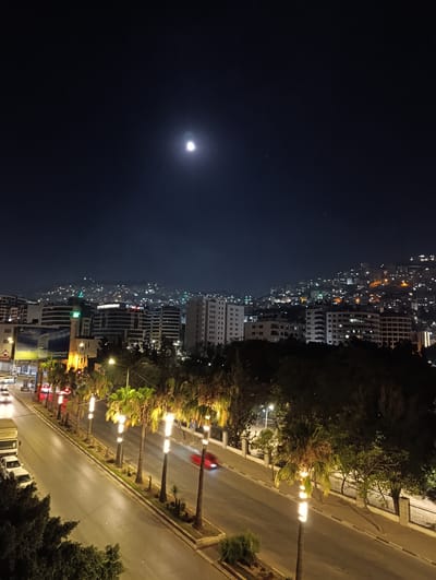 The image depicts a nighttime view of a city, likely Nablus, Palestine, from an elevated perspective. The sky is dark blue with a visible crescent moon and some faint stars. In the background, the cityscape is illuminated by numerous lights, suggesting a densely populated area, with buildings cascading up a hillside. In the mid-ground, several modern buildings with lit windows and distinct architectural features are visible, including one with a green-lit minaret, possibly a mosque. In the foreground, a street is lined with palm trees that are illuminated by upward-facing lights, creating a dramatic effect. Several cars are present on the road, with one red car in motion leaving a motion blur, and a white truck and car parked on the side. The street is paved and has sidewalks. Trees and foliage are also present, contributing to the urban environment. The overall impression is one of a vibrant city at night.