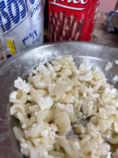 This close-up image, taken in Krivodol, Bulgaria, features a metal bowl filled with a mixture of food, flanked by two different packaged items.

The main subject is the food in the shiny metal bowl, which consists of small, pale yellow, curved pasta shapes (resembling macaroni) mixed thoroughly with white, crumbly cheese, similar to cottage cheese or feta. The texture suggests a soft, prepared dish, possibly macaroni with cheese, that is ready to be consumed or further prepared.

To the left of the bowl, a vibrant red cylindrical packaging is partially visible. It displays the characteristic "Coca" script, indicating the Coca-Cola brand, and features an image of several brown chocolate-filled wafer sticks or biscuit rolls.

Below the red packaging, a white and blue plastic bag or wrapper is also partially seen. Visible text includes "energ dran," likely an abbreviation for "energy drink," and "product" on a small yellow label. The bag features a pattern of interconnected blue shapes, possibly an abstract representation of energy or a stylized logo.

The scene appears to be an indoor setting, likely a kitchen or dining area, with casual lighting. There is no specific activity depicted other than the arrangement of these items, suggesting a paused moment before a meal or snack. The overall impression is one of a simple, domestic food preparation or mealtime setting.
