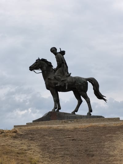 The image shows a dark bronze equestrian statue of a soldier with a rifle slung over his shoulder, mounted on a horse. The statue is positioned on a concrete pedestal, which is itself set upon a tiered, stone-faced embankment. The background consists of a cloudy sky. The ground in the foreground is rough and covered with dry, brown grass and dirt. The statue is located on a hilltop, with a view of the sky and what appears to be a natural landscape. The style of the statue and the clothing of the soldier suggest a military historical context, potentially related to Russian history.
