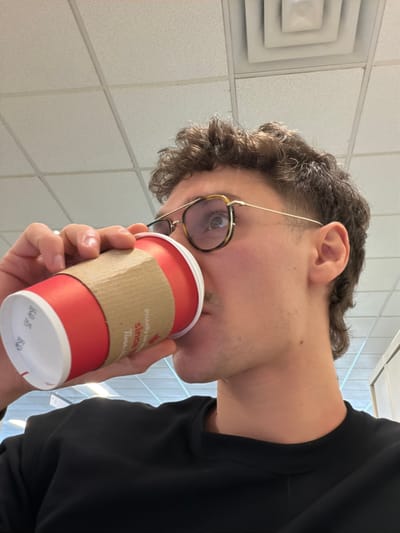 A male individual is depicted in a close-up, profile view, drinking from a red Tim Hortons disposable cup within an indoor commercial or institutional setting in Mirabel, Canada. The subject has short, curly brown hair styled in a short mullet, a light mustache, and wears gold-framed eyeglasses with tortoiseshell accents. His right hand grips the cup, which features a beige corrugated sleeve and a white lid. The background consists of a white drop ceiling with visible grid lines, acoustical panels, and a multi-layered square air vent.