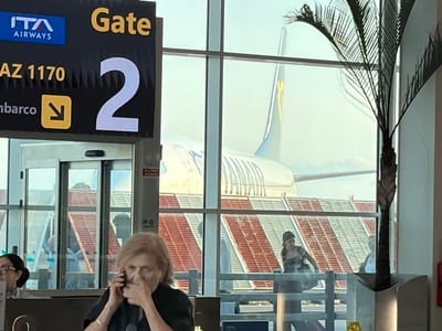 This media file depicts an indoor scene at an airport, likely a gate area, with a view of an airplane parked outside. A dark sign with yellow lettering indicates "Gate 2" and "AZ 1170". The ITA Airways logo is also visible on the sign.

In the foreground, a woman is talking on her cell phone and appears to be touching her nose. To her left, another woman with glasses is partially visible. Through the large windows, an airplane's tail is prominently displayed, featuring the yellow and blue colors of what appears to be a Ryanair aircraft based on the tail design, although the text is not fully legible. A palm tree is also visible, partially obscuring the view of the aircraft.

Behind the aircraft, a red and white tiled roof structure is visible, suggesting the airport might be in a region with this type of architecture. The lighting suggests it is daytime, with bright ambient light filtering through the windows. The overall atmosphere seems to be that of a typical airport waiting area, with people preparing to board a flight. The presence of an airplane and gate information clearly indicates travel is about to occur.