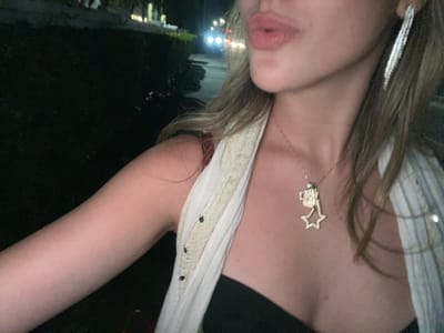 A woman poses with her head tilted, wearing a white top with embellishments along the strap. A necklace with a star and charm hangs around her neck. The background is dark, hinting at an outdoor setting with distant lights suggesting streetlights or a cityscape. Her facial expression is relaxed, with slightly pursed lips.