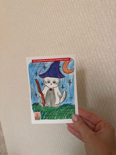 The image shows a drawing of a white cat dressed as a wizard. The cat is wearing a blue pointed hat and holding a brown stick. It has a crescent moon and stars in the background, with green grass at the bottom. Text at the top of the drawing reads "ПРИ ПОДДЕРЖКЕ РОССИЙСКИХ ЖЕЛЕЗНЫХ ДОРОГ" (With the support of Russian Railways). Below this, it says "Богданова Дарья, 10 лет" (Bogdanova Darya, 10 years old) and "Казань" (Kazan), followed by the title "Кот-волшебник" (Wizard cat). A small, colorful emblem is visible at the bottom left corner of the drawing. A hand with painted nails is holding the drawing against a textured, beige background. The location of Кирпичного завода № 1, Russia is not depicted in this image.