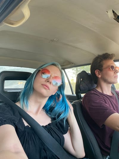 The image depicts two individuals, a woman and a man, inside a car. The woman, with vibrant blue hair, is in the foreground, wearing sunglasses with red and blue lenses and a black top. She is leaning slightly towards the camera, with her right arm resting on her lap. The man, with brown hair, is in the driver's seat, wearing sunglasses and a burgundy t-shirt. He is looking forward, seemingly focused on driving. The car's interior is visible, including the ceiling, headrests, and seatbelts. The outdoor environment outside the car windows suggests a daytime scene, possibly during sunset given the lighting. The location is identified as Калыс-Ордо жаңы курулушу, Kyrgyzstan.