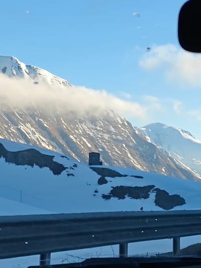 The image shows a landscape of snow-covered mountains under a partly cloudy sky. In the foreground, a metal guardrail is visible, with a small bird perched on it. A dark, square structure is centered on a snow-covered ridge. Behind the ridge, mountains rise steeply, partially obscured by clouds, with patches of snow. The image is likely taken from inside a vehicle due to reflections on what could be the car window. Based on the prompt information, the location is in the city of Ganisi, Georgia.