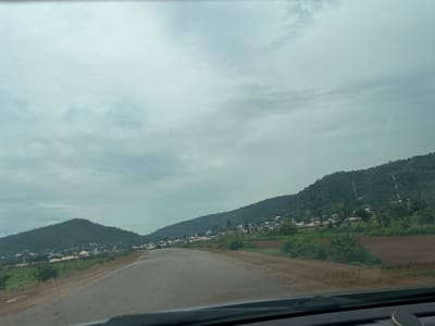 The image depicts a scenic view of a road winding through a landscape with lush green hills and a small village. The sky is overcast with a cloudy, hazy appearance, suggesting it might be early morning or a cloudy day. The road is paved and appears to be a two-lane highway, with a dirt shoulder on the right side.
The village is nestled at the foot of the hills, characterized by numerous buildings with various roof types, including what appear to be corrugated metal and tiled roofs. Some of the structures seem to be residential homes, while others might be public buildings like schools or community centers. There are also several communication towers visible on the slopes of the hills.
The vegetation is dense, with trees and bushes covering the hillsides. In the foreground, there are patches of tilled earth, indicating agricultural activity. A few vehicles are seen on the road, including cars and what looks like a truck, suggesting the road is active with local traffic.
The overall impression is a serene, rural setting in Nigeria, possibly Koton Karifi, given the context. The cloudy weather contributes to a soft, diffused light, creating a calm and tranquil atmosphere. There are no visible people in the image, and no text is present. The scene captures a typical view from a moving vehicle, looking out towards the surrounding countryside.