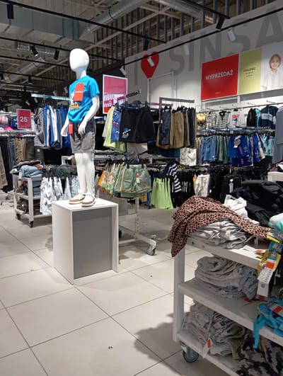 The image shows a retail display of children's clothing in what appears to be a store in Wołomin, Poland. The setting is a well-lit retail environment with white tiled floors and industrial-style ceiling elements.
The main subjects are clothing items displayed on racks and shelves, featuring various garments like shorts, t-shirts, and pants. A prominent feature is a child-sized mannequin wearing a blue t-shirt with a graphic design and grey shorts, positioned to showcase the apparel. Nearby, a sign with the text "WYPRZEDAŻ" (Polish for "SALE") in red is visible, indicating a promotional event. Another sign displays "25" followed by a punctuation mark, likely indicating a price or discount.
The overall activity is the presentation of merchandise for sale. There are no discernible people actively shopping in this frame, but the context clearly implies a commercial setting. The lighting suggests daytime, and there are no clues about the weather. The arrangement of clothing and display units is typical of a clothing store, with a focus on children's wear. The brand "SONIC" is visible on some of the clothing items.