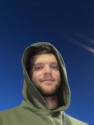 A man with fair skin, a beard, and a hooded sweatshirt is looking directly at the camera. The background appears to be a dark blue, possibly a night sky. The photo is taken from a low angle, looking upwards at the man. The photo does not contain enough information to place this image in Springfield, United States of America.