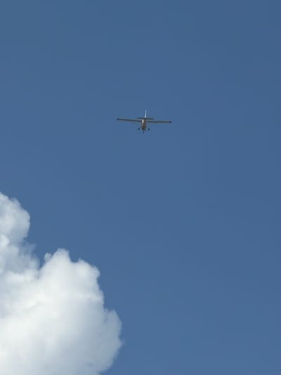 A small, light-colored airplane is flying in a clear blue sky. The plane is small and appears to be a propeller-driven aircraft. Its wings are extended horizontally, and its tail is visible. In the lower left portion of the image, a large, fluffy white cloud is present. The airplane is positioned in the upper center of the frame. There are no people visible in the image. The scene appears to be an outdoor environment with a clear sky and some clouds. The image does not provide any specific details about its location in Lebanon, United States of America.