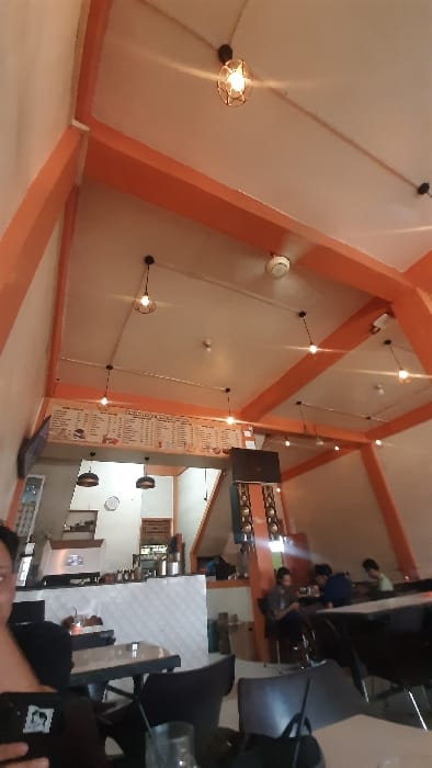 The image displays the interior of a cafe or casual restaurant in Manetunong, Indonesia. The venue features a high, vaulted white ceiling with prominent orange structural beams forming an angular design. Multiple pendant light fixtures, each containing an exposed filament bulb within a wire cage, are suspended from the ceiling, complemented by smaller recessed lights and white smoke detectors.
A large, white menu board displaying text and illustrations is mounted high on the back wall, extending across a service area. Below the menu board, a white-tiled counter with a dark countertop holds various beverage preparation equipment and containers. A circular wall clock is visible on the wall behind this counter.
In the lower left foreground, a person's left hand and a portion of their upper body, dressed in dark clothing, are visible, holding a smartphone. A clear glass with a dark straw sits on a light-colored table in front of this individual. The dining area is furnished with dark tables and chairs. In the mid-ground to the right, three male individuals are seated at a table. One male, in a dark shirt, looks downwards. Another male, wearing a blue shirt, also looks down. A third male is partially visible further behind them. They appear to be occupied with personal devices or engaged in quiet conversation. To the right of the service counter, a tall stack of black audio speakers with silver accents is positioned, flanked by orange structural elements. The interior relies primarily on artificial lighting.