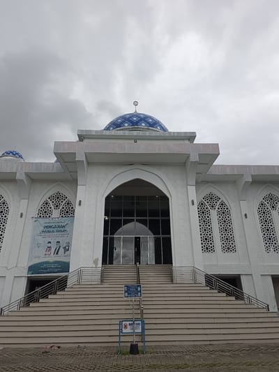 The image shows the exterior of a mosque, likely the Islamic Center Attaqarrub in Pidie Jaya Regency, Indonesia. The mosque is a white building with ornate white relief patterns on its facade and intricately designed windows with Islamic geometric patterns. A prominent blue-domed roof with a crescent moon finial crowns the central part of the structure.

In front of the mosque, a wide set of stairs leads up to the entrance. Two metal railings flank the stairs, and at the bottom of the stairs, there are signs indicating "TEMPAT WUDHU PRIA" (Wudu place for men) and "TEMPAT WUDHU WANITA" (Wudu place for women), as well as a "BATAS SUCI" (Sacred boundary) sign instructing to leave sandals and shoes. The ground in front of the stairs is paved with interlocking blocks.

A banner is visible hanging on the left side of the building, above the stairs. Although partially obscured, text in Indonesian can be read, including "Hadiri dan Ikuti....!" (Attend and Follow...!) and "PENGAJIAN MAJELIS TAKLIM" (Majlis Taklim Recitation). Below this, the name of the Islamic Center and the regency are mentioned.

The sky is overcast and cloudy, suggesting either early morning or late afternoon, or simply a cloudy day. The overall atmosphere is solemn and suggests a place of religious gathering. The scene captures the architectural beauty of the mosque and its functional elements for worshippers.