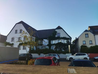The image shows a street scene in Leverkusen, Germany, featuring a white, multi-story building with a dark tiled roof that appears to be a historical structure, possibly a guesthouse or seminar house given the signage visible. The building is partially covered in green vines. In front of the building, several cars are parked along the street, including a black SUV, a silver station wagon, a red hatchback, and a white SUV. A large, grey truck trailer is also visible on the left side of the frame. The sky is clear and blue, suggesting it is daytime. The foreground is dominated by a low concrete wall, behind which the cars and building are situated.