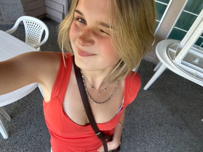 A woman with blonde hair, wearing an orange dress and a brown strap across her chest, is taking a selfie from a low angle. She is winking at the camera. She is wearing jewelry, including a necklace and a nose ring. In the background, there are white plastic chairs and a building with windows. The surface under the chairs appears to be concrete. The location may be Kelowna, Canada, but there are no identifiable landmarks to confirm this.