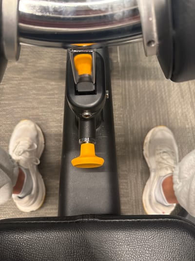 An overhead shot reveals gym equipment: a black bench with a yellow adjustment knob on its side. Parts of the padded seat are visible, along with a shiny metal support. Two feet in white sneakers, with white socks, are standing on the grey patterned floor around the equipment. The focus is primarily on the adjustment mechanism and surrounding structural details.