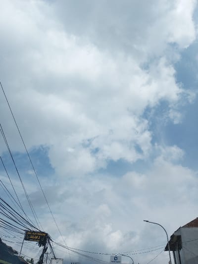 The image presents an upward-angled view of a daytime sky with partial cloud cover, observed from an urban or semi-urban environment in Cibadak, Sukabumi Regency, Indonesia. The upper two-thirds of the frame are dominated by a blue sky with scattered white cumulus clouds.

In the lower left quadrant, a dense cluster of black utility wires, likely electrical or communication, crisscrosses horizontally and diagonally across the frame, originating from an obscured pole structure. Attached to these wires and visible in the lower-left center is a black rectangular sign with white text, partially obscured, which reads "JUAL ALAT PANCING JALAN 2" (Sell Fishing Gear Road 2) and, below that, "KAB. SUKABUMI" (Sukabumi Regency) along with a partial phone number.

Towards the lower center-right, a smaller rectangular blue and white sign, possibly displaying "PUSAT RADIAL" or similar text with a logo, is visible. Adjacent to it on the far right, a modern-style street lamp with a curved arm and an LED fixture extends upwards. The corner of a building with a light-colored wall and a red-tiled roof is visible on the extreme right edge of the frame. No individuals or discernible actions are present within the scene.