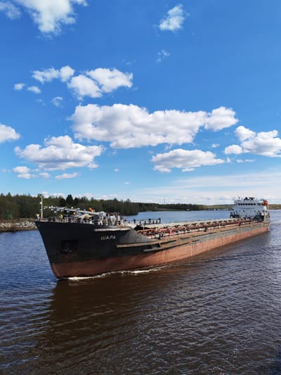 The image depicts a large, rusted cargo ship, identified as "ШАЛА" (SHALA), moving along a river. The ship is heavily laden and is positioned towards the left of the frame, angled slightly away from the viewer. The river's water is dark and reflective, showing ripples from the ship's movement. The backdrop features a line of dense green trees marking the shoreline. The sky is bright blue with scattered, puffy white clouds. The location is likely Ivanov Bor, Russia, based on the Cyrillic script on the ship and the general environment. There are no people visible in the image, only the cargo ship and its surroundings. The action is the ship navigating the river.