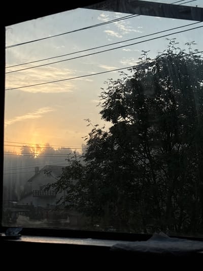 The image depicts a view through a window of a rural landscape at dawn. The sun is rising, casting a warm orange glow on the sky and illuminating a patch of mist. Power lines crisscross the sky, and a large, dark tree dominates the right side of the frame. In the distance, a house with a chimney and a balcony is visible, along with several small outbuildings. The overall impression is one of a peaceful morning in a village setting. The location is identified as Jāņupe, Latvia.