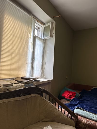 In a dimly lit room, a worn sofa with a textured beige fabric is positioned in the foreground. To the right, a bed is covered with a blue sleeping bag and a red shirt is draped over a green pillow. The walls are painted a muted olive green. A window, partially obscured by translucent white material that appears to be taped or adhered to the glass, lets in faint light, casting horizontal shadows. Several books and newspapers are stacked on a windowsill. A simple, dark-colored wooden chair is visible next to the sofa. The scene suggests a modest living space within a dwelling in посёлок опытного хозяйства «Ермолино», Russia.