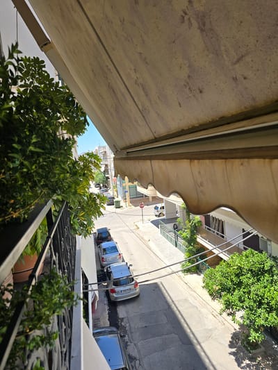 The image shows a view from a balcony in Ilion, Greece, looking down at a street lined with parked cars and buildings. A green leafy plant is in the foreground on the left, partially obscuring a black metal railing. An awning extends from the building above, casting a shadow. The street is paved, with a few cars parked along the right side. One car, a silver hatchback, is prominent in the lower portion of the image. Further down the street, more cars are visible, as well as a green dumpster. Buildings with balconies and windows line the street. A stop sign is visible on the right side of the street in the distance. The sky is clear and blue.