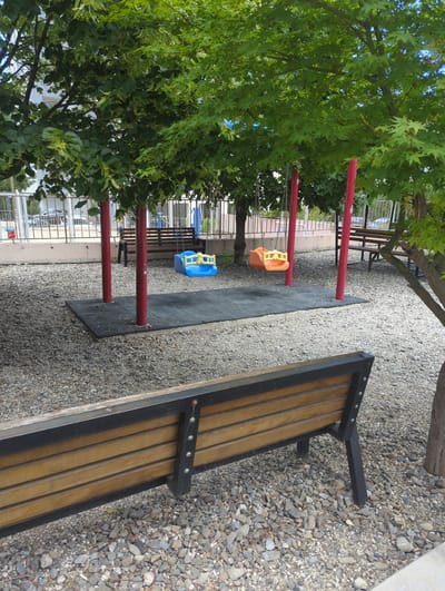 The image depicts a playground area in Tbilisi, Georgia, characterized by a gravel ground surface and lush green trees providing shade. In the foreground, a wooden bench with a black metal frame is positioned, facing away from the camera. Behind the bench, a swing set structure with red support poles is visible. Two swings are attached: one is a blue plastic swing with a yellow frame, and the other is an orange plastic swing also with a yellow frame. These swings are suspended over a black rubberized mat, likely for safety. Further in the background, other benches and playground equipment can be seen, along with a metal fence and vehicles parked in the distance. The overall environment suggests an outdoor recreational space, possibly part of a school or park in Tbilisi.