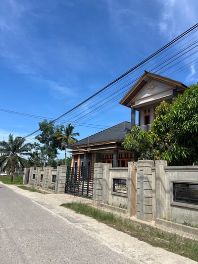 The image captures a residential building in Dalam, Indonesia, on a bright, clear day. The building appears to be multi-storied, with a distinct triangular pediment featuring a circular emblem. Electrical wires run diagonally across the frame, partially obscuring the blue sky, which has a few wispy clouds. The building's exterior is a mix of light-colored concrete, dark roofing, and wooden accents around windows and on balconies. A concrete fence with decorative elements and a black metal gate encloses the property. Lush green trees, including a palm tree, are visible around the house, suggesting a tropical environment. The scene is viewed from the roadside, with an asphalt road and a concrete curb taking up the lower right portion of the image. The overall impression is that of a modern home in a sunny, possibly rural or suburban, Indonesian setting. No people are visible in the frame, and there is no discernible activity taking place. The lighting suggests it is daytime, likely late morning or early afternoon, given the brightness and the sharp shadows. There is a small red marking with the number "677" on the concrete fence, but no other visible text.