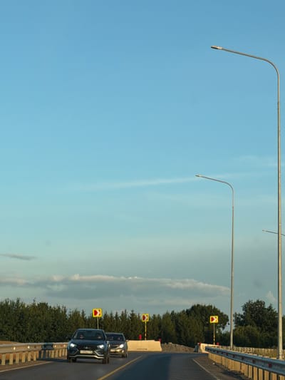 This media file captures a road scene in Derzhavino, Russia. Two cars are visible on a paved road, with one car in the foreground and another behind it. The foreground car is a dark SUV, and its headlights are on. The car behind it appears to be a lighter-colored sedan, also with its headlights illuminated.

The road is bordered by a concrete barrier with reflective markers. Along the roadside, there are several yellow and red directional signs indicating curves in the road. Tall, green trees line the far side of the road. Above, the sky is a soft blue with a few wispy white clouds, suggesting it might be late afternoon or early evening. Two tall streetlights with curved arms extend over the road.

The scene appears calm and ordinary, with no obvious signs of distress or unusual activity. The lighting suggests it's not midday, and the weather seems clear. The visible text on the license plate of the foreground car is partially obscured but appears to be in Cyrillic script, consistent with the specified Russian location.