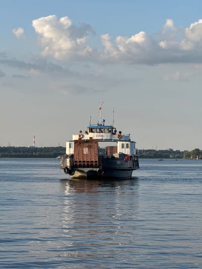 A ferry boat is moving away from the camera on a wide, calm river under a blue sky with scattered white clouds. The ferry is dark gray with a white upper structure. On the back of the ferry, there is a large, brown cargo container. There are also several people visible on the deck of the ferry. In the distance, a tree-lined shore is visible, with some industrial structures, including a tall red and white smokestack, on the left side. The water has ripples and reflects the sunlight. The ferry's hull has several black tires along its side. The name "СПН8" is visible on the side of the ferry. This scene is consistent with the location of Verhnij Uslon, Russia, where ferries operate across the Volga River.