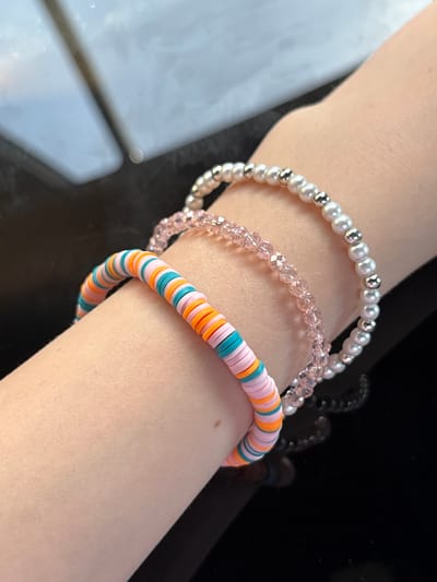 This is a close-up, high-angle, eye-level shot of three bracelets on a person's arm. The bracelets are stacked on top of each other and are resting on a dark, reflective surface. The top bracelet is made of small, white, pearl-like beads interspersed with small, silver beads. The middle bracelet is made of small, faceted, pink beads. The bottom bracelet is a colorful, flat disc bead bracelet with alternating colors of pink, orange, and teal. The background consists of a light blue, out-of-focus area, likely representing a ceiling or light source. The image does not contain any information about the city of Tresserre, France.