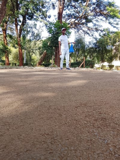A single adult male is depicted standing on a dark grey-brown asphalt surface in Kikuyu Township ward, Kenya. The paved surface, characterized by embedded small stones, dominates the lower two-thirds of the image. The male, positioned slightly left of center, wears a light grey short-sleeved t-shirt, light grey trousers, dark blue athletic shoes, and a baseball cap with a red and white brim. He holds a crumpled blue plastic bag in his extended left hand, while his right arm rests at his side. The background consists of dense green foliage, featuring several large trees with thick, reddish-brown trunks and numerous smaller bushes, under bright daylight conditions. No other individuals or significant objects are present.