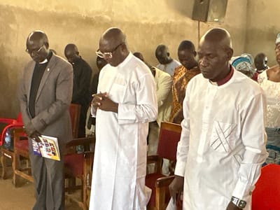 The media file shows a group of men, likely clergy, gathered indoors, possibly in a church or religious hall in Kufana, Nigeria. They are all dressed in formal attire. The individuals in the foreground are bowing their heads and have their hands clasped, suggesting they are engaged in prayer or a moment of reflection.

One man on the left is wearing a grey suit with a clerical collar, holding a booklet in his hand. To his right, another man is dressed in a white robe, his hands clasped in front of him. Further to the right, a third man, also in a white robe with a clerical collar, has his eyes closed. Other men in the background are similarly attired and appear to be participating in the same activity. The scene is illuminated by overhead lighting, creating a warm, indoor ambiance. The backdrop consists of a textured, light-colored wall. In the background, parts of wooden chairs with red upholstery are visible.

There is no indication of the time of day or weather conditions. The atmosphere appears solemn and respectful. The overall impression is one of a religious gathering or service. No visible text is discernible in the media file.