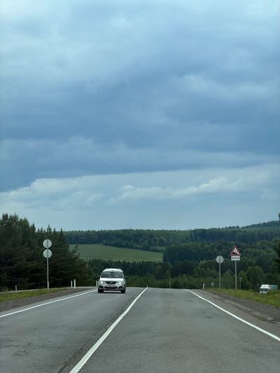 The image depicts a wet asphalt road curving through a rural landscape under a cloudy sky. The perspective is from a vehicle driving on the road, looking ahead and slightly to the side. A white car is visible in the distance, traveling in the same direction. The road is bordered by lush green trees, forming a dense forest on one side. In the background, rolling hills with fields of green are visible, suggesting a countryside setting.

On the side of the road, there are traffic signs. One prominent sign is a triangular warning sign with a red border, indicating a steep descent of 5% and a distance of 700 meters. Another sign features two circular white discs on a pole, the purpose of which is unclear from the image. In the far distance, a white truck is partially visible.

The weather appears overcast, with a bright, diffused light, suggesting it is daytime, likely late morning or afternoon. The wetness of the road indicates recent rain or ongoing light precipitation. The overall mood is serene and natural, highlighting the beauty of the rural scenery. The location is identified as Vakhrino, Russia. There is visible text on the license plate of the white car, which reads "M600XH".