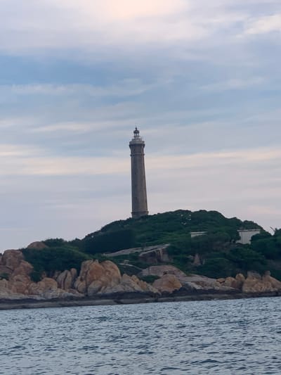 A tall, slender lighthouse stands on a rocky, tree-covered hill overlooking a body of water. The lighthouse is made of stone and has a white lantern room at its peak. The hill is covered in lush green vegetation and dotted with large, weathered rocks. The water in the foreground is a deep blue, with gentle ripples. The sky above is overcast with a soft, diffused light, suggesting it may be dawn or dusk. The scene appears to be a coastal landscape, possibly a remote island or a rugged shoreline.