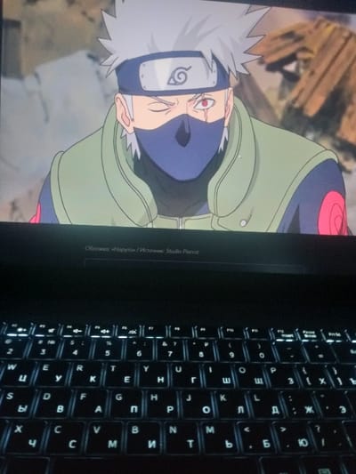 A screen displays an image of Kakashi Hatake from Naruto. He wears his signature headband and mask. Below the image are the Russian words "Обложка: «Наруток / Источник: Studio Pierrot," indicating the cover image's origin. To the right of the screen is a backlit laptop keyboard featuring both English and Russian letters. The keyboard is dark, with the white backlight illuminating the keys.