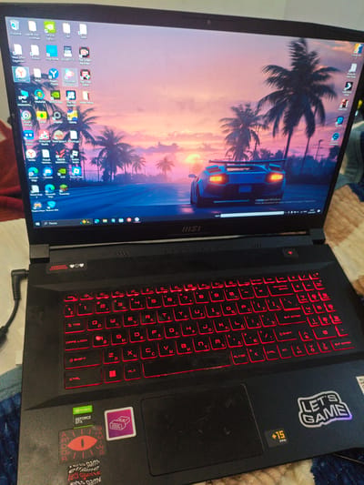 The image shows a black MSI gaming laptop with a vibrant wallpaper of a futuristic car driving down a palm-lined road at sunset. The laptop screen displays various application icons, including "NVIDIA GeForce," "Roblox," "Discord," and several Yandex applications. The keyboard has red backlighting, and several stickers are visible on the laptop's body. One sticker says "LET'S GAME" with "+15 FPS" below it. Another sticker features an eye symbol and "ARDOR GAMING." The laptop is placed on a patterned surface, likely a bed or couch. The image is taken from a slightly elevated, frontal perspective. There are no people visible in the image, and the environment is indoors. The context of Kalach-na-Donu, Russia, is not depicted or implied by the visual information in the image.