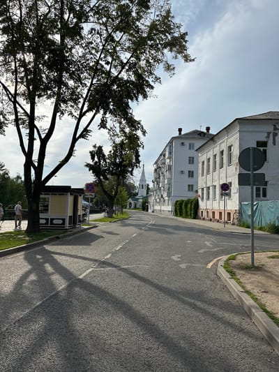 The image depicts a street scene in Tolbukhino, Russia, under a partly cloudy sky. A paved road occupies the foreground, with prominent shadows of trees cast across it from the left. On the left side of the road, a green grassy area is visible, bordered by a curb. Beside the grass is a small, light yellow kiosk with a dark roof and trim. Two individuals, a man and a woman, are walking on the grassy area near the kiosk. Further down the road, a white spire of a church can be seen in the distance. To the right of the road, there are several buildings. The closest is a white, two-story building with a dark roof, followed by a taller, white apartment building. A dark green vine covers a portion of the wall between these buildings. Road signs are present on the right side of the street, including a circular sign with a red border and a blue center, indicating no stopping or parking. The environment appears to be a town or city setting with a mix of residential buildings and public spaces.