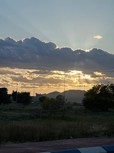 The image depicts a sunset scene in Sant Vicent del Raspeig, Spain. A mountain silhouette is visible in the distance, with street lights and trees lining the horizon. The sky features a combination of dark, heavily textured clouds on the left, contrasting with streaks of golden sunlight breaking through thinner cloud layers towards the center. The sun's rays create a layered effect, illuminating portions of the landscape. The foreground shows a strip of green vegetation, with a corner of a structure or barrier visible in the bottom right. The orientation of the image is portrait, but rotated 90 degrees counter-clockwise.