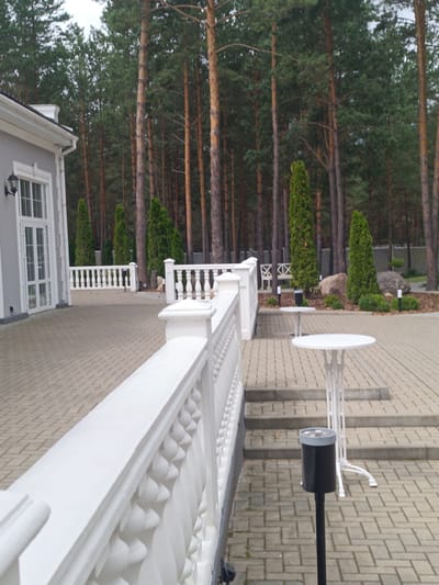 This is an outdoor scene on a paved patio area, likely a restaurant or event venue, located in the Старобінскі сельскі Савет, Belarus. The patio is paved with rectangular interlocking pavers and features a decorative white balustrade along a raised section. In the background, a dense forest of tall pine trees dominates the landscape under a cloudy sky.
Several white outdoor tables are visible on the patio. One round table with an ornate base is in the foreground on the right, and another similar table is slightly further back and to the left. A dark cylindrical ash stand on a pole is positioned near the stairs leading up to the patio from the lower paved area. A white railing separates the upper patio from a lower paved area. On the left side of the image, part of a building with a grey facade, white trim, and large glass doors with white frames is visible.
The environment is designed for outdoor seating and relaxation, with the natural backdrop of the pine forest suggesting a semi-rural or park-like setting. There are no people visible in the image.
