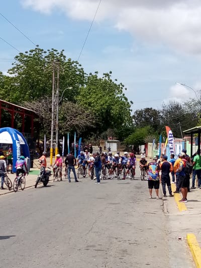 The image shows a cycling race in Guatacaral, Venezuela. A group of cyclists in racing attire is lined up on a street, ready for the start. Several people stand along the side of the road, observing the race. There is a blue tent with white lettering visible on the left side of the image. A motorcycle is parked near the cyclists. The road is asphalt, and there is a yellow curb on the right side. Buildings and trees are visible in the background. Banners are also present near the cyclists. The sky is partly cloudy.