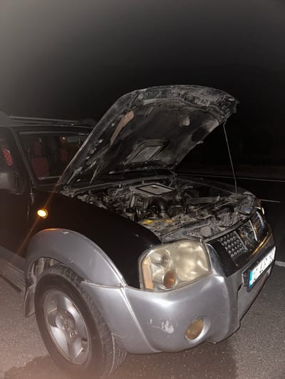 A silver and black car with its hood open sits on a street at night. The front right tire is visible, as is the engine underneath the open hood. The license plate is attached to the front of the car. The photo was reportedly taken in Ula, Turkey, although there are no indicators of this in the image itself. The door to the car appears to be open, and red material is visible inside. There are no people visible. The environment is dark, suggesting a nighttime setting.