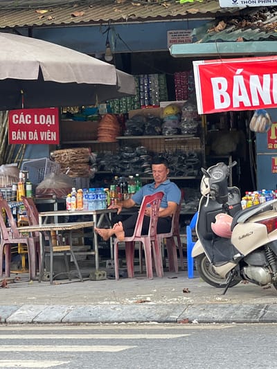 A middle-aged Vietnamese man with short, dark hair is sitting on a plastic chair at a table outside a shop in Tân Quang Commune, Vietnam. He is wearing a blue polo shirt and dark pants, with his bare feet resting on the seat of another chair. The shop appears to be selling various goods, with shelves of products visible in the background. A red sign with white text that reads "CÁC LOẠI BÁN ĐÁ VIÊN" (meaning "Types of ice cubes for sale") is prominently displayed. A red and white banner with the word "BÁ" visible is also hanging from the shop. A light-colored scooter is parked to the right of the man, with a pink helmet resting on its front. The scene is outdoors, with a sidewalk in the foreground and a street with a crosswalk visible. The shop has a corrugated metal roof.