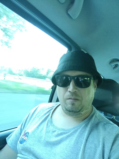 A man wearing a black bucket hat and sunglasses is taking a selfie in a car. He is wearing a gray t-shirt with a NASA logo on the left chest. The man has light skin and stubble on his chin. The car's interior is visible, including the headliner and a portion of the passenger seat. Outside the car window, trees and a bright sky are visible, suggesting an outdoor daytime scene. The location is identified as Psáře, Czechia.