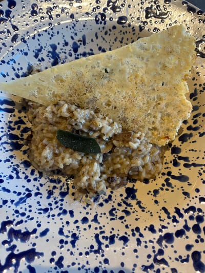 The image displays a close-up, top-down view of a single portion of food served on a round, white ceramic plate adorned with an irregular blue splatter pattern. The central element of the dish is a creamy, light brown risotto, exhibiting individual grains of rice and darker, amorphous inclusions. A single, dark green, oval-shaped leaf, consistent with sage, is positioned on top of the risotto. Partially embedded into the risotto and extending upwards is a large, golden-yellow, triangular crisp object with a thin, porous, and somewhat irregular texture, consistent with a cheese frico, lightly speckled with black particles, likely ground pepper. No individuals or specific actions are depicted. The image does not contain any visual information to verify its location in Dorchester, United Kingdom.