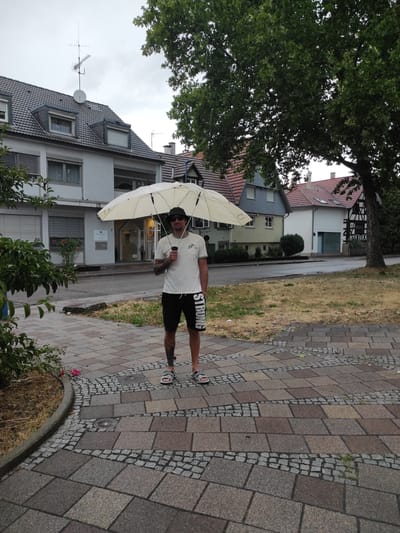 A man is standing on a paved walkway in front of a building in Waldbronn, Germany, holding an open, cream-colored umbrella. He is wearing sunglasses, a white t-shirt, and black shorts. The man has tattoos on his left leg. In the background, there are several residential buildings with pitched roofs, some with visible chimneys. A large tree with green leaves is positioned to the right of the frame, partially obscuring the sky and buildings. The walkway is made of interlocking rectangular stones and smaller cobblestones. There is a patch of dry grass to the left of the walkway, and a pink rose is visible near the curb. The sky is overcast.