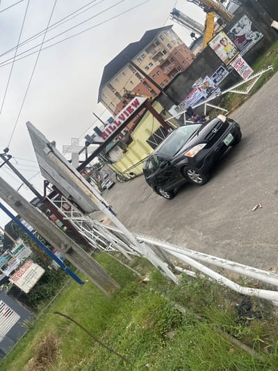 A black car is parked on a street beside a sidewalk, with a building and a "Hillview" sign behind. Several posters and construction cranes are visible nearby. On the other side of the sidewalk, there is a grassy area. Some power lines and advertisements can be seen in the background.
