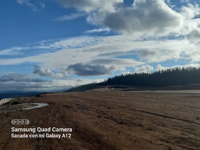 The image depicts an expansive outdoor landscape in Santa Juana, Chile, under a partly cloudy sky. The scene is dominated by a vast expanse of reddish-brown, disturbed earth, indicative of cleared land or a dirt track, with faint tire marks visible across its surface. On the far left foreground, a light-colored wooden fence runs along a downward slope, adjacent to several small, irregular puddles of standing water in ground depressions. The midground to background features a dense, dark line of coniferous trees forming a distinct horizon along the right side. To the left, the land slopes down towards distant, hazy hills or mountains. The sky, occupying the upper half of the image, displays a mix of bright blue patches and numerous white to grey cumulus clouds, with sunlight illuminating the scene. No individuals or animals are visible. A white text overlay in the bottom-left corner states "Samsung Quad Camera Sacada con mi Galaxy A12".